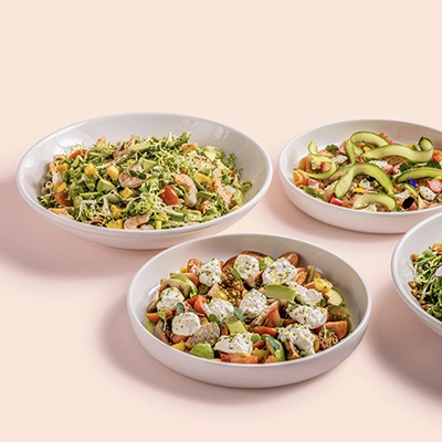 Three Elio salad bowls with fresh vegetables, avocado and toppings on a pink background