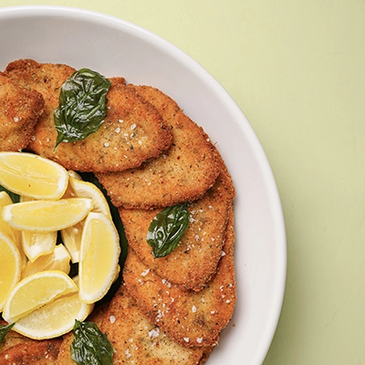 Breaded chicken escalope in a white bowl with lemon slices and fresh basil on a sage green background