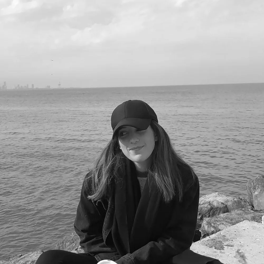 Black and white picture of Yoland wearing a cap smiling with the ocean behind her